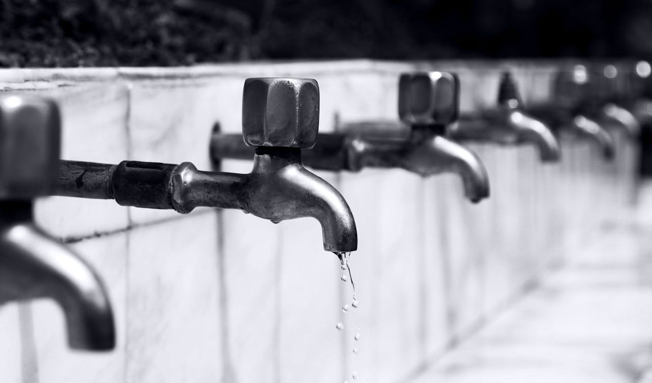 about-us-02 Black and white image of a row of faucets with water dripping, highlighting plumbing details.